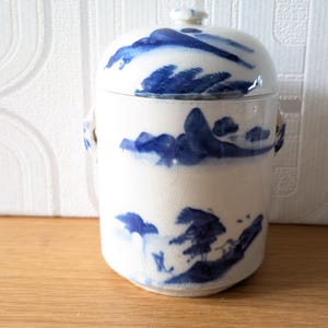 May include: A white ceramic jar with a lid, adorned with blue landscape designs. The jar has a cylindrical shape with small handles on the sides. The lid features a small knob. The design is reminiscent of traditional Asian pottery.