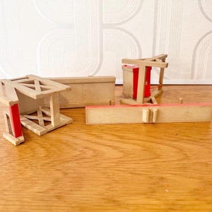 May include: A collection of miniature wooden furniture pieces including a bench and chair frames with light tan wood grain and bright red accents. The pieces feature cross-braced supports and rectangular panels, with some components lying flat on the surface.