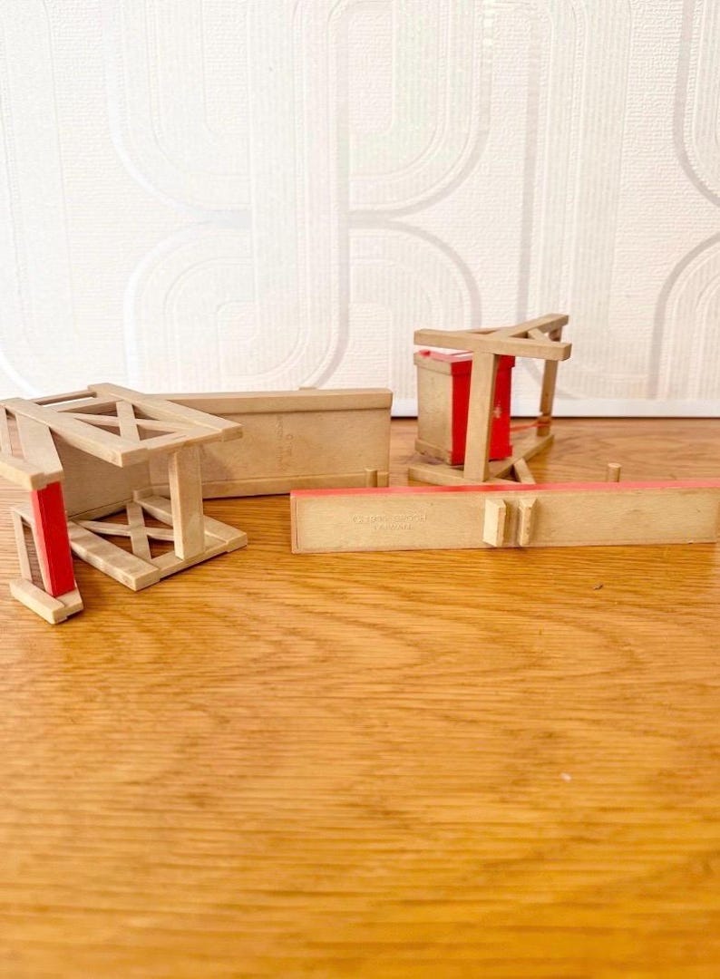 May include: A collection of miniature wooden furniture pieces including a bench and chair frames with light tan wood grain and bright red accents. The pieces feature cross-braced supports and rectangular panels, with some components lying flat on the surface.
