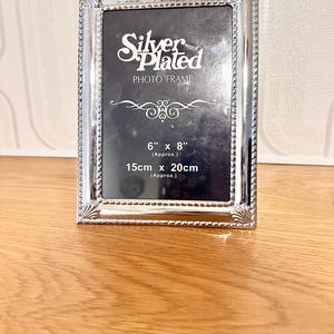 May include: A silver-plated photo frame with a rectangular shape. The frame has a decorative beaded edge and holds a photo space of approximately 15cm x 20cm. The text "Silver Plated PHOTO FRAME" is displayed on the black background.