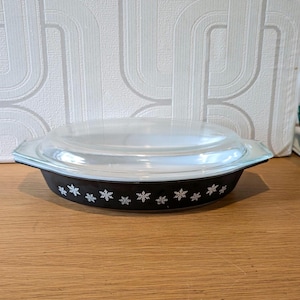 May include: Black oval casserole dish with a clear glass lid. The dish has a white snowflake pattern around the rim. The dish is on a wooden surface.