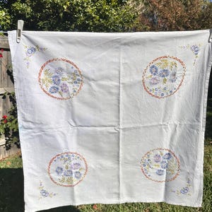 May include: A square white fabric cloth featuring four circular embroidered motifs containing blue and purple floral patterns with orange borders. Additional small floral sprigs are embroidered in each of the four corners of the white textile.