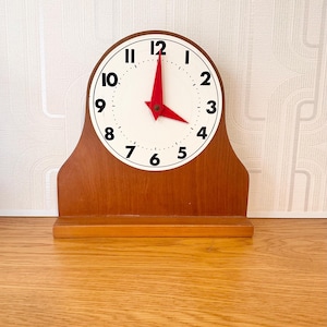 May include: A wooden mantel clock with a white face and black numerals. The clock hands are red. The clock is set on a wooden surface. The clock's design is a classic, vintage style.