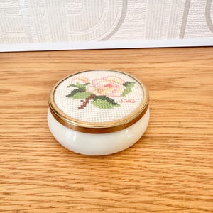 May include: A round white ceramic trinket box with a brass-toned metal rim. The lid features a cross-stitched floral design of a pink and yellow flower with green leaves on white fabric, with the initials JWG embroidered in pink on the right side.