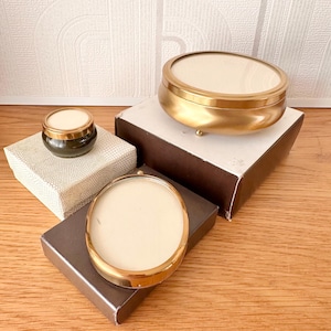 May include: A collection of three circular containers with metallic gold rims and off-white lids. One small dark green glass jar sits on a textured white box, while two larger containers with gold bases rest on brown boxes, displaying a polished finish.