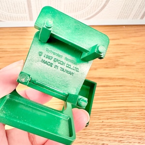 May include: A green plastic miniature chair with four legs and a flat seat. The underside features molded text reading Sylvanian families, 1987 EPOCH CO., LTD., and MADE IN TAIWAN. The object is constructed from solid green material with a smooth finish.
