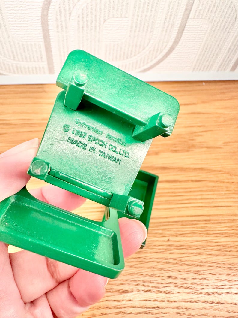 May include: A green plastic miniature chair with four legs and a flat seat. The underside features molded text reading Sylvanian families, 1987 EPOCH CO., LTD., and MADE IN TAIWAN. The object is constructed from solid green material with a smooth finish.