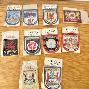May include: A collection of souvenir badges from various regions, including Cornwall, Devon, Somerset, and Dorset. Each badge features a unique design with text indicating the location. The badges are in their original packaging.