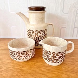 May include: A vintage ceramic coffee set featuring a coffee pot with a lid, a creamer, and a sugar bowl. The set has a speckled beige finish with brown floral patterns. The coffee pot is approximately 8 inches tall.