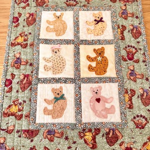 May include: A rectangular quilted wall hanging featuring a central grid of six squares, each displaying a unique fabric teddy bear appliqué. The border is a sage green fabric patterned with small teddy bears, surrounded by a daisy-print frame.