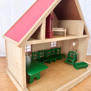 May include: A two-story wooden dollhouse with a bright pink roof. The ground floor features a green dining table, four green chairs, a green high chair, and a small green cradle. The upper floor contains a tall dark red grandfather clock.