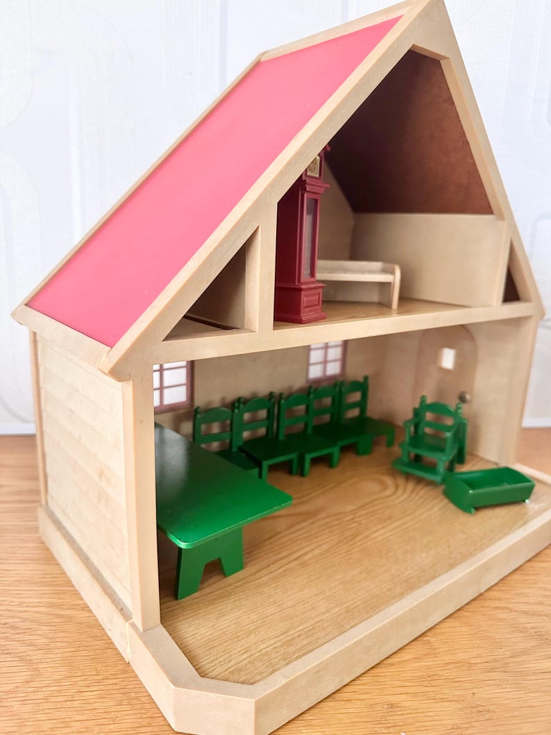May include: A two-story wooden dollhouse with a bright pink roof. The ground floor features a green dining table, four green chairs, a green high chair, and a small green cradle. The upper floor contains a tall dark red grandfather clock.