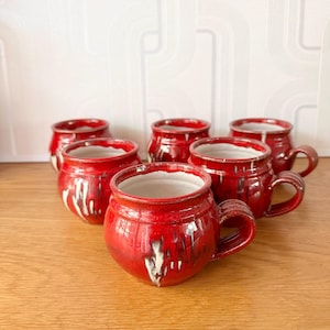 May include: Six red ceramic mugs with white drip glaze detailing. Each mug features a rounded body, a handle, and a white interior. The mugs are displayed on a wooden surface.
