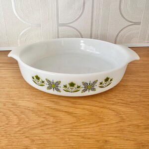 May include: White oval casserole dish with a green and yellow floral pattern around the rim. The dish has two handles. It is on a wooden surface. The background is a light-coloured wallpaper.