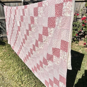 May include: A rectangular quilt featuring a patchwork design of alternating dusty rose and cream-colored fabric squares. The pattern consists of various floral, lattice, and vine prints arranged in a diagonal grid, finished with a solid cream border.