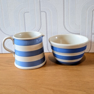May include: A small white pitcher and bowl set with blue horizontal stripes. The pitcher has a handle and a spout. The bowl is round. Both pieces are on a wooden surface.