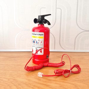 May include: A red fire extinguisher with a black handle and nozzle. A red coiled cord with a clear connector is attached. The extinguisher has a label with instructions. The items are on a wooden surface.