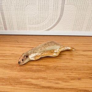 May include: A ceramic sculpture of a small, elongated animal, possibly a rodent or weasel, with a light brown and cream color scheme. The sculpture is lying on a wooden surface. The animal has a detailed head and body.