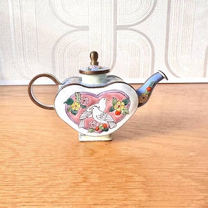 May include: A heart-shaped teapot featuring a white enamel finish with a central pink heart design containing two white birds surrounded by colourful flowers. The teapot has a brass handle, a brass lid knob, and a light blue spout with floral accents.