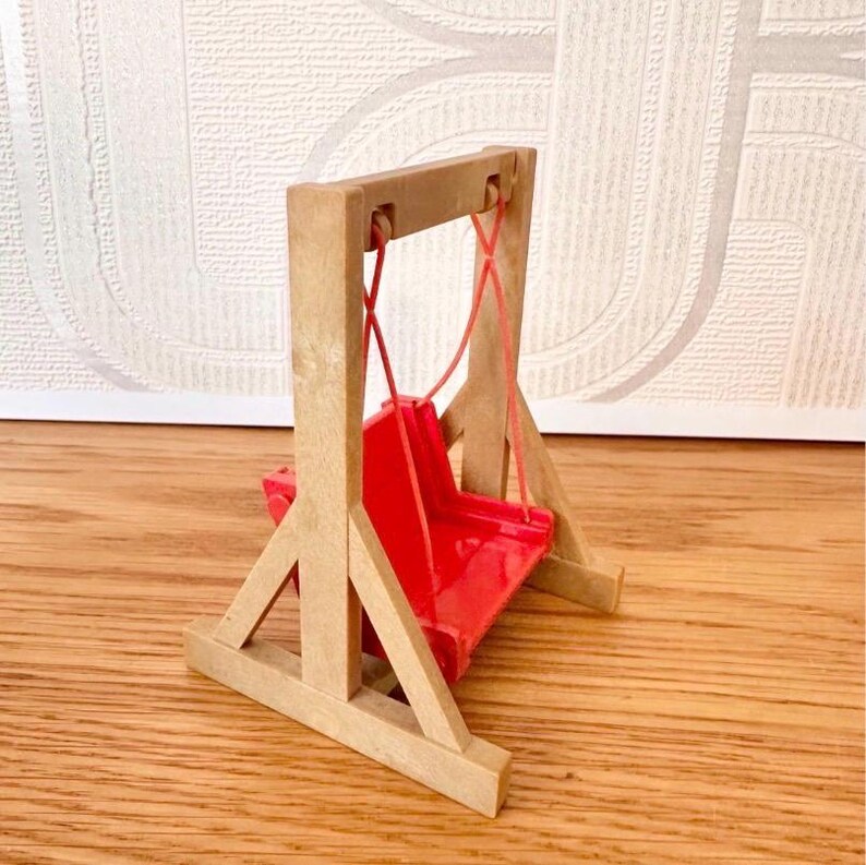 May include: A miniature toy swing set featuring a light brown wooden frame with a triangular base and a bright red plastic seat suspended by thin red cords. The structure is designed with two vertical posts and diagonal supports for stability.