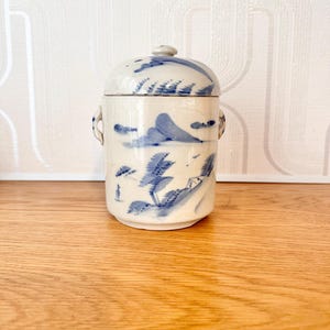 May include: A ceramic jar with a lid, featuring a blue and white landscape design. The jar has a cylindrical shape with small handles and a rounded lid. The landscape design includes mountains, trees, and clouds. The jar is approximately 6 inches tall.