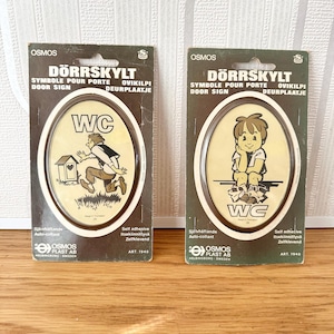 May include: Two vintage self-adhesive door signs with oval illustrations. One shows a person running towards a small house, the other a child sitting. Both signs have the letters "WC" and text in multiple languages, including "Door Sign".