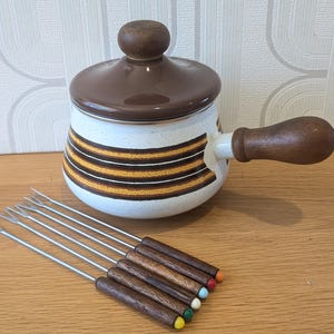 Peut inclure: Un caquelon à fondue vintage avec un couvercle et une poignée marron, et un corps blanc avec des rayures marron et orange. Comprend six fourchettes à fondue avec des manches en bois et des embouts colorés. Un ensemble de cuisine rétro.