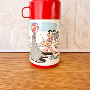 以下が含まれることがあります： 赤と白のヴィンテージ魔法瓶。赤い蓋とハンドルが付いています。魔法瓶には、プルートとアザラシがビーチボールで遊んでいる漫画のイラストが描かれています。ベースは白で、赤い縁取りがあります。