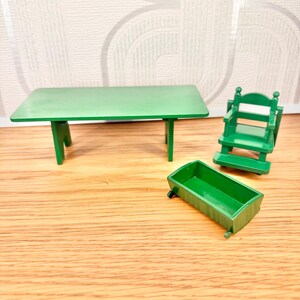 May include: A miniature green furniture set consisting of a rectangular table with four legs, a high-backed chair with armrests, and a small rectangular open-top container. All items are made of solid green plastic with a smooth, uniform finish.