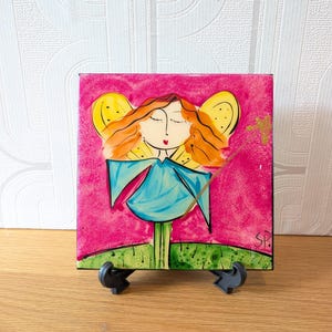 May include: A square decorative tile featuring a stylised illustration of a fairy with orange hair, yellow wings, and a blue dress, holding a gold wand against a vibrant pink background. The fairy stands on a green grassy base with the initials SP.