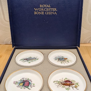 May include: A boxed set of four Royal Worcester Bone China coasters. Each coaster has a different seashell design with gold edging. The navy blue box has gold lettering.
