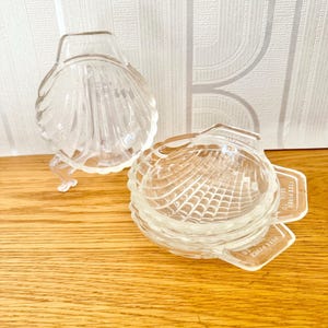 May include: Two clear glass seashell-shaped dishes with a textured surface. The dishes are made by Kella Bean and say "Aged Sherry Made to"