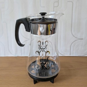 May include: A glass coffee pot with a black handle and a chrome lid. The pot has a black and white design on the side and sits on a black stand with three legs.