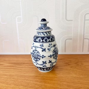May include: A small, white ceramic jar with a lid, decorated with blue floral and geometric patterns. The jar has a bulbous body and a rounded lid with a small, dark blue knob. The design is reminiscent of traditional Asian pottery.