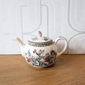 May include: A cream-coloured ceramic teapot featuring a colourful floral pattern with pink, red, and blue blossoms on green stems. The lid and upper rim are decorated with a black and white geometric border pattern, and the lid has a small knob.