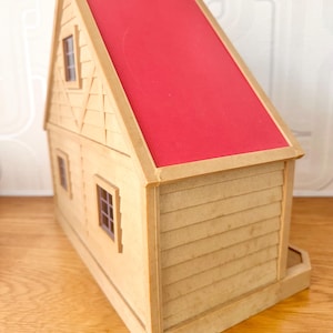 May include: A toy house with tan walls and a bright red roof. The structure features horizontal siding on the lower level, a gabled roofline, and two small windows with dark frames visible on the side wall. The exterior has a textured finish.