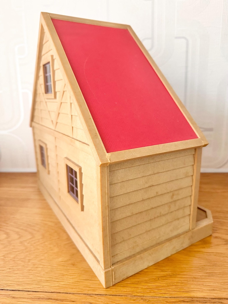 May include: A toy house with tan walls and a bright red roof. The structure features horizontal siding on the lower level, a gabled roofline, and two small windows with dark frames visible on the side wall. The exterior has a textured finish.