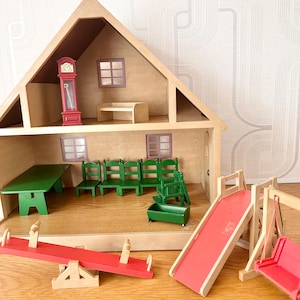 May include: A wooden dollhouse with two levels, featuring a red grandfather clock, a green table, four green chairs, and a green cradle. Outside the house are a red seesaw, a red slide, and a red swing set, all constructed from wood.