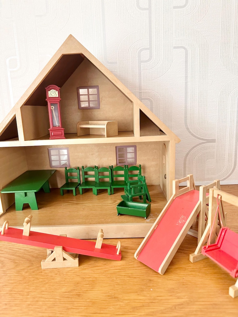 May include: A wooden dollhouse with two levels, featuring a red grandfather clock, a green table, four green chairs, and a green cradle. Outside the house are a red seesaw, a red slide, and a red swing set, all constructed from wood.