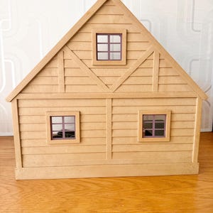 May include: A tan-colored toy house with a triangular roof, horizontal siding, and three square windows with dark purple frames. The structure features a central vertical beam and diagonal accents on the upper level, creating a simple, symmetrical facade.