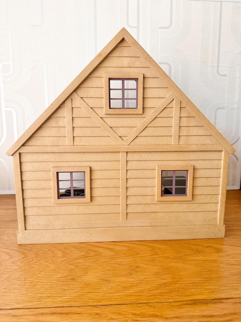 May include: A tan-colored toy house with a triangular roof, horizontal siding, and three square windows with dark purple frames. The structure features a central vertical beam and diagonal accents on the upper level, creating a simple, symmetrical facade.