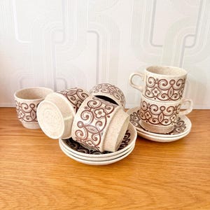 May include: A set of vintage ceramic coffee cups and saucers. The cups and saucers are beige with a brown floral pattern. The cups have handles and are stacked. The saucers are round.
