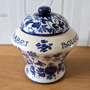 May include: A ceramic jar with a lid, featuring a blue and white floral design. The jar has a bulbous shape with a flared base and a rounded lid. The word "ABBRI" and "BOLOG" are written on the jar.