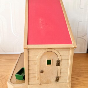 May include: A beige toy house with a bright pink slanted roof, featuring a single arched door with a small square window, metal hinges, and a round knob. A small green rectangular container sits on the attached beige base platform.