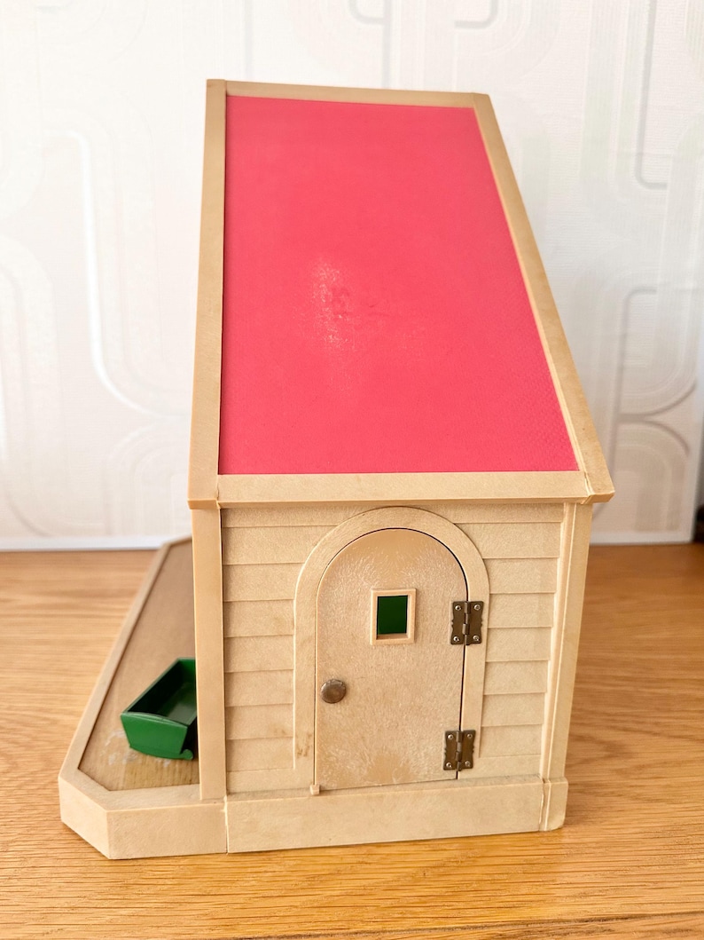 May include: A beige toy house with a bright pink slanted roof, featuring a single arched door with a small square window, metal hinges, and a round knob. A small green rectangular container sits on the attached beige base platform.