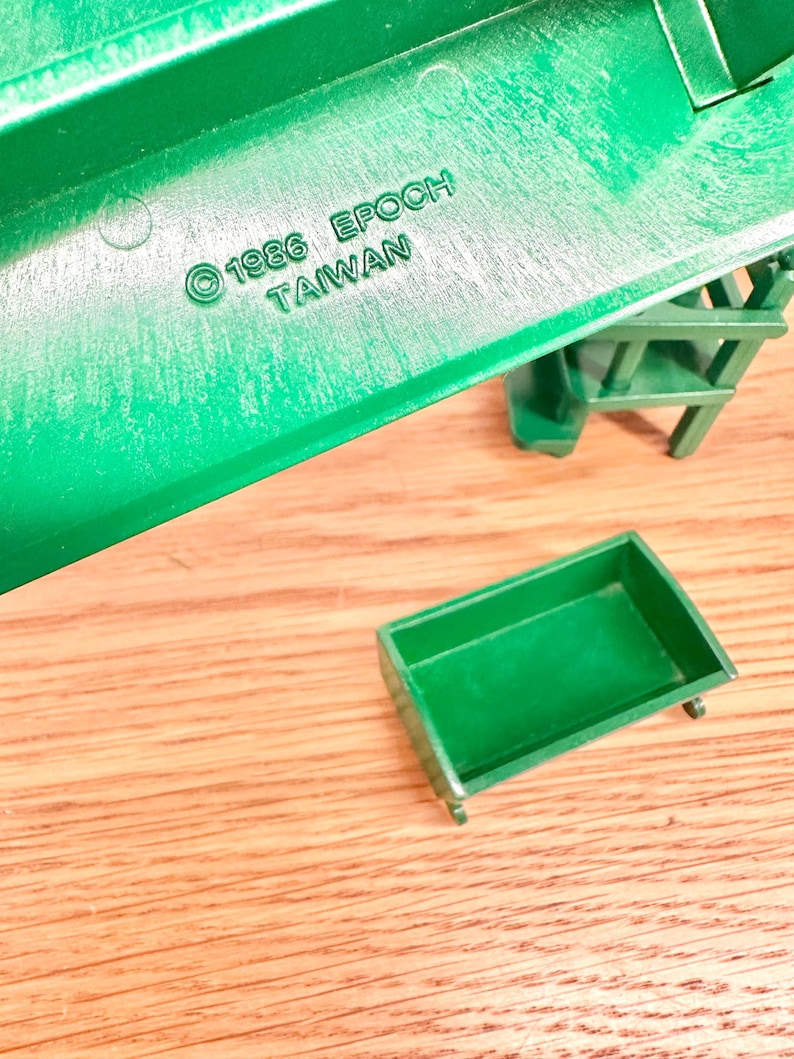 May include: A green plastic toy structure featuring a small rectangular green bin and a staircase component. The structure is embossed with the text 1986 EPOCH TAIWAN. The pieces are molded from solid green plastic with a smooth finish and visible structural details.
