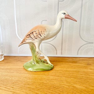 May include: A ceramic figurine of a white bird with brown markings standing on a green base. The bird has a long, thin beak and is posed with its head turned to the side.