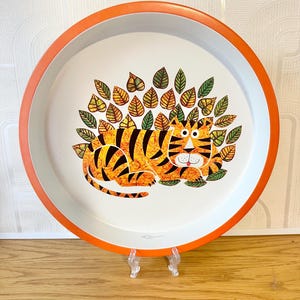 May include: A circular serving tray with an orange border and a white interior. It features a cartoon tiger design in orange and black, surrounded by green and yellow leaves. The tray is displayed on a clear stand.