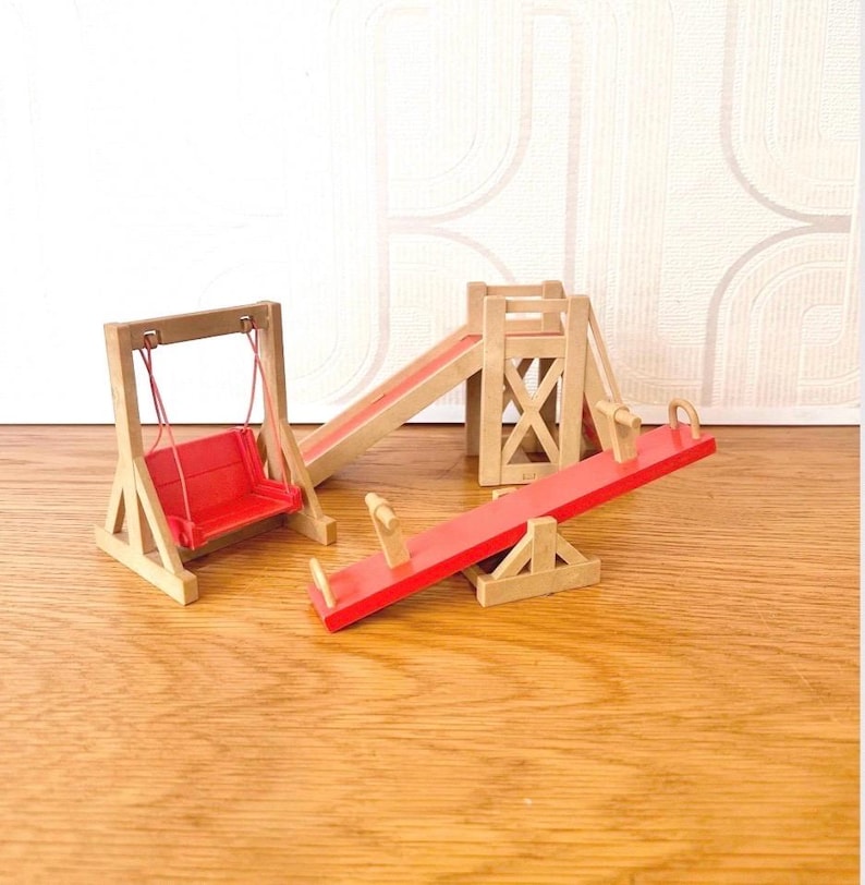 May include: A miniature wooden playground set featuring a swing with a red seat, a slide with a red ramp, and a seesaw with a red board. The structures are constructed from light-colored wood and include red plastic or painted components.