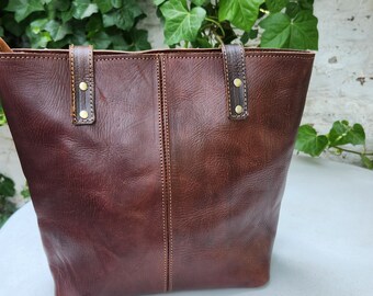 Puede incluir: Un bolso tote de cuero marrón con dos asas y detalles dorados. El bolso tiene un diseño cosido y está hecho de cuero texturizado.